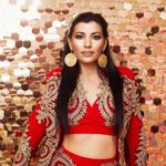 Attractive female wearing red costume and round earrings looking at camera while standing in light studio on background with golden sequins