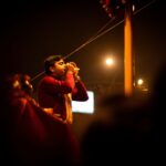 A young man participates in a traditional religious ceremony at night in Varanasi, India, blowing a conch shell with devotion.