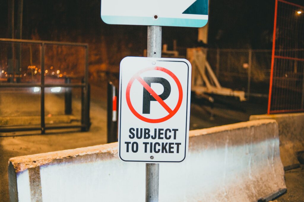 Prohibition parking road sign with Subject To Ticket inscription placed on street of city near concrete barrier in evening time