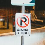 Prohibition parking road sign with Subject To Ticket inscription placed on street of city near concrete barrier in evening time