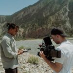 Two filmmakers capture scenic mountain river with professional camera.