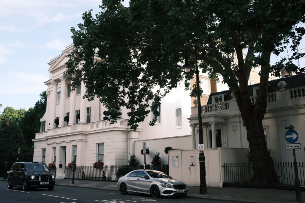 Charming white townhouse facade on a sunny street with parked cars and lush trees.
