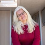 A cheerful woman with glasses and white hair smiling warmly inside a cozy camper van.