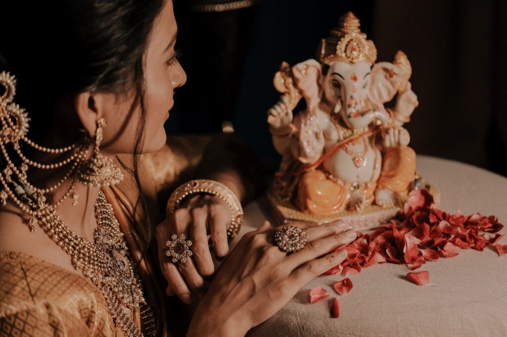 A woman artistically dressed in jewelry admires a Lord Ganesha idol surrounded by rose petals.