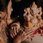 A woman artistically dressed in jewelry admires a Lord Ganesha idol surrounded by rose petals.