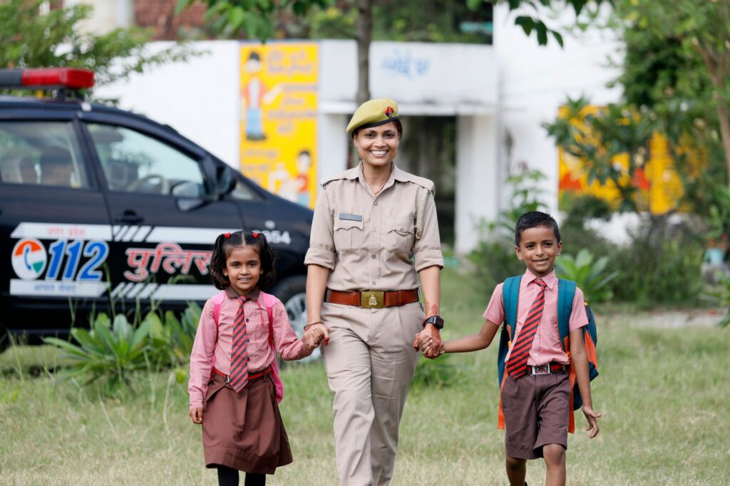 A smiling police officer walks with two children in school uniforms holding hands outdoors.