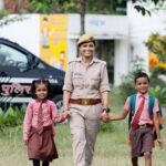 A smiling police officer walks with two children in school uniforms holding hands outdoors.