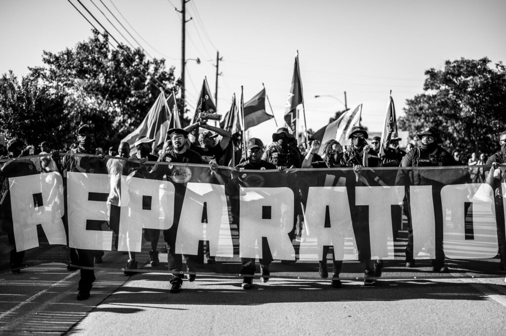 A group of people marching for reparations holding a large banner on a city street.