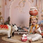 Joyful child in traditional attire with festive decor celebrating Janmashtami indoors.