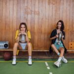 Two young women sitting on a bench with a trophy and a boombox, showcasing fashion in a sports setting.