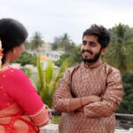 A smiling Indian couple in traditional attire, standing on a balcony, surrounded by lush greenery.