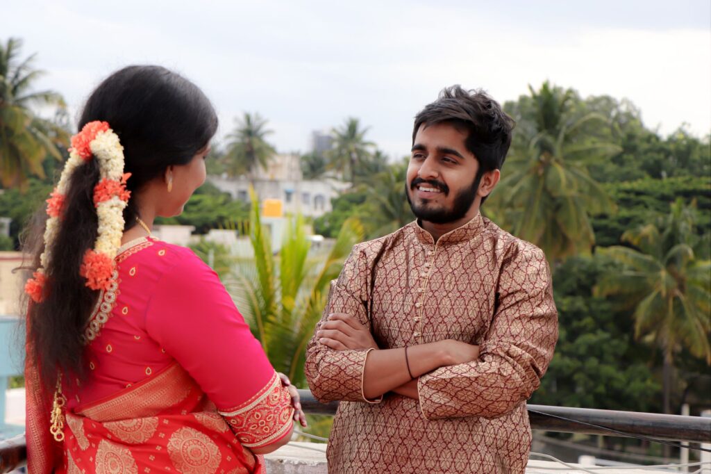 A smiling Indian couple in traditional attire, standing on a balcony, surrounded by lush greenery.