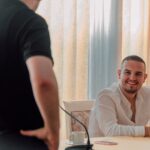 Two men in a casual business meeting setting. One is smiling while seated, communicating friendly interaction.