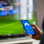 Close-up of a person holding a smartphone with a VPN app, streaming sports on TV.