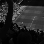 Fans cheering during a high-energy soccer match with players on the field, captured in black and white.