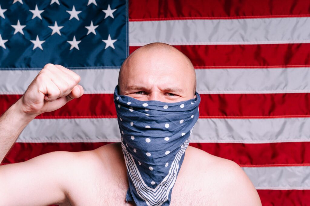 Bald man wearing a bandana as a face mask with American flag backdrop, raising his fist.