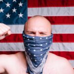 Bald man wearing a bandana as a face mask with American flag backdrop, raising his fist.
