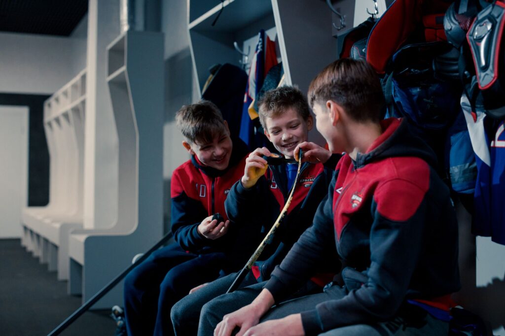 Three young boys in hockey gear taping sticks and laughing in a locker room.