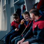 Three young boys in hockey gear taping sticks and laughing in a locker room.