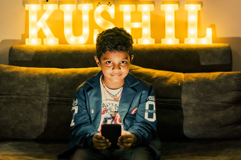 A young boy sits indoors holding a smartphone with illuminated KUSHIL letters in the background.