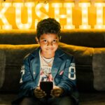 A young boy sits indoors holding a smartphone with illuminated KUSHIL letters in the background.