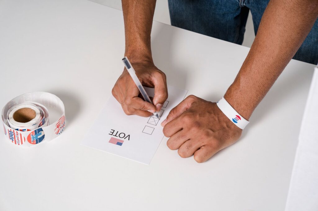 A person wearing a wristband fills out a voting ballot with a pen. Voting stickers visible.