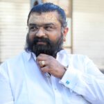Portrait of a confident adult man with a beard and spectacles, wearing a white shirt outdoors.