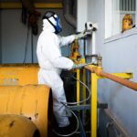 Worker in full protective suit and gas mask handles industrial pipes indoors in Indonesia.