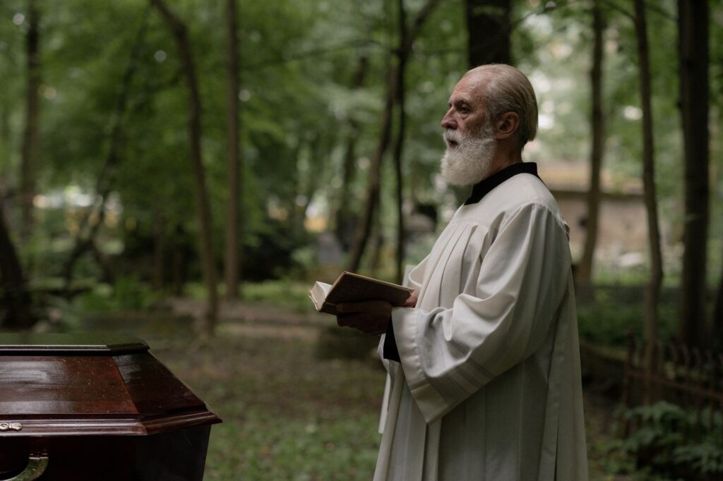 A priest dressed in ceremonial robes holds a holy book during an outdoor funeral in a forest setting.