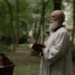 A priest dressed in ceremonial robes holds a holy book during an outdoor funeral in a forest setting.