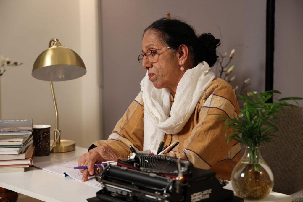 An elderly woman sits at her desk writing with a typewriter in a cozy indoor setting.
