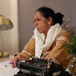 An elderly woman sits at her desk writing with a typewriter in a cozy indoor setting.