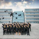 Group of graduates celebrating and tossing caps outside modern building.