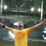 A badminton player in a yellow shirt celebrates triumphantly in an indoor stadium.