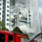 Firefighters respond to a building fire with smoke and emergency vehicles in an urban area.