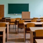An empty classroom featuring wooden desks, chairs, and a large green chalkboard.