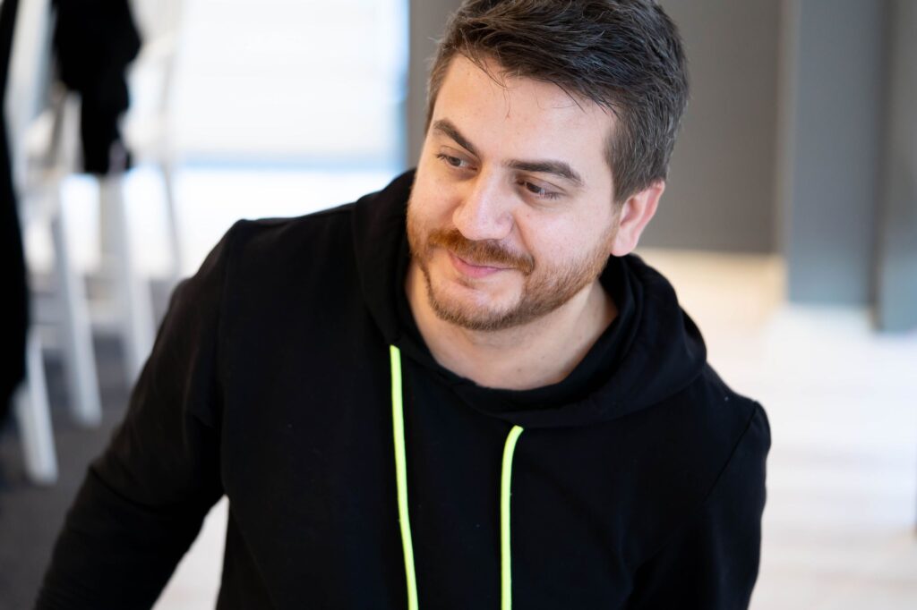 Close-up of a man wearing a black hoodie, looking off-camera with a smile.
