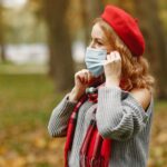 A woman in a red beret adjusts her face mask in a park during fall.