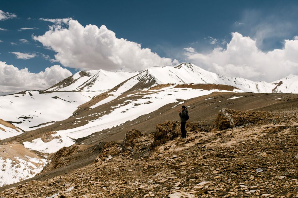 Side view of distant anonymous tourist standing on slope of hill against snowy mountain peaks and cloudy sky in highlands in winter day