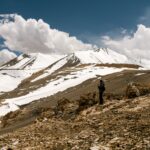 Side view of distant anonymous tourist standing on slope of hill against snowy mountain peaks and cloudy sky in highlands in winter day