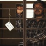 Man standing behind bars holding a sign asking 'May I Go Out?' Indoor setting with dramatic tone.