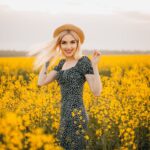 Woman in sundress and hat smiling amidst vibrant yellow flowers in a sunny field.