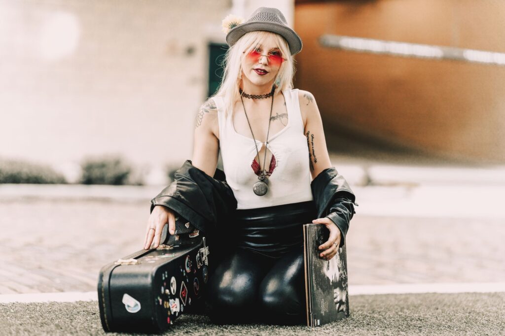 A modern musician with a guitar case, wearing trendy clothes and accessories, sitting outdoors.