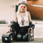 A modern musician with a guitar case, wearing trendy clothes and accessories, sitting outdoors.