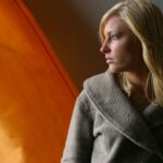 A thoughtful woman in warm light gazes outside, conveying introspection.
