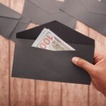 A hand holds a black envelope containing a 100 dollar bill on a wooden surface.