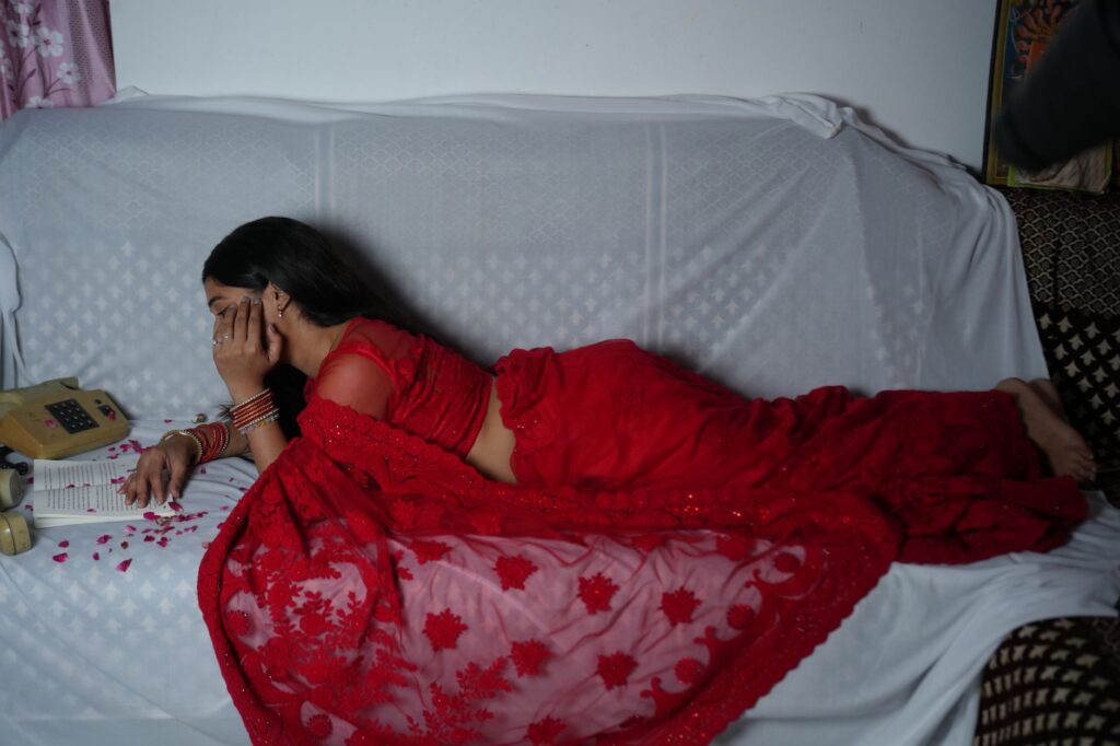A woman in a vibrant red saree lies on a couch, lost in thought or reading indoors.