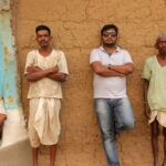 Four men standing and sitting against a mud wall in Balaghat, India.