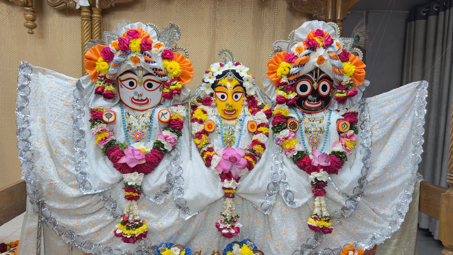 Vibrant Jagannath, Balabhadra, and Subhadra idols adorned with flowers and ornaments indoors.