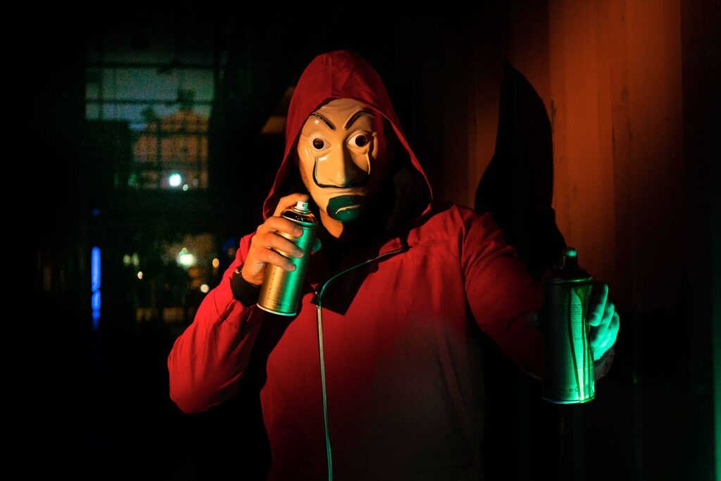 A masked person in a red hoodie holds spray paints, creating street art at night.